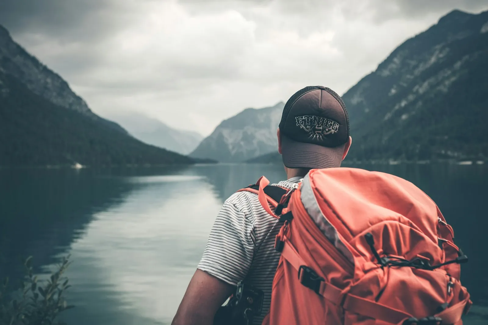 A traveler with an orange backpack looking out over a mountain lake