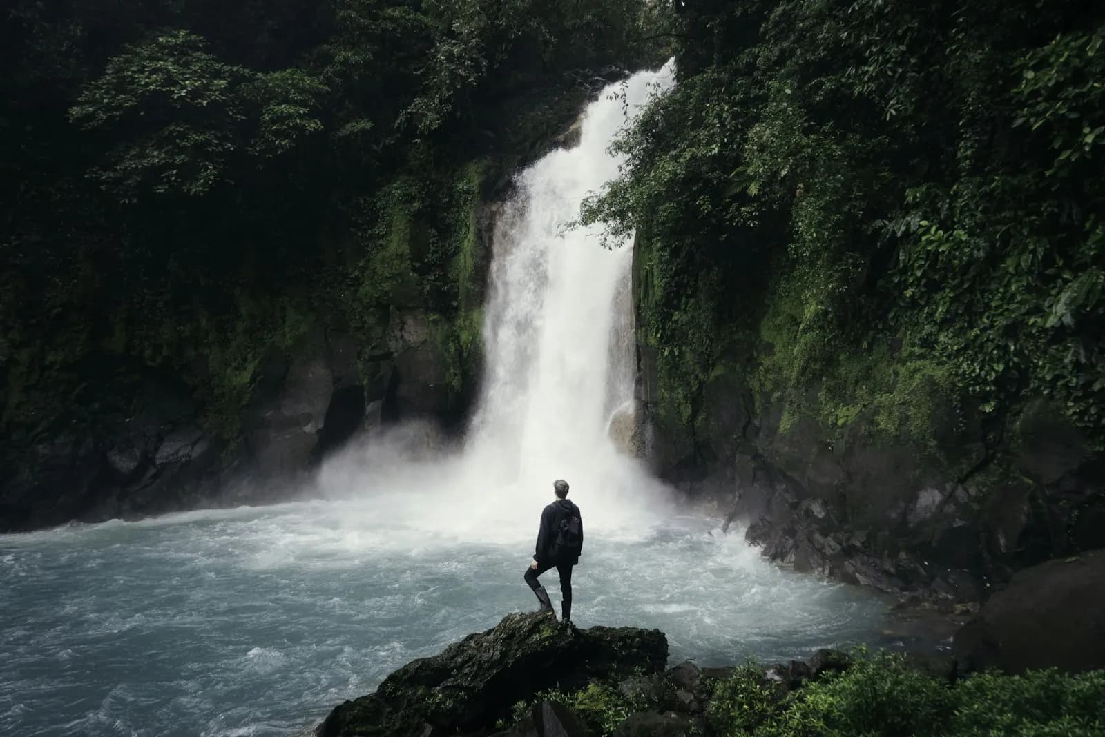 A traveler standing on a rock in front of a tall waterfall